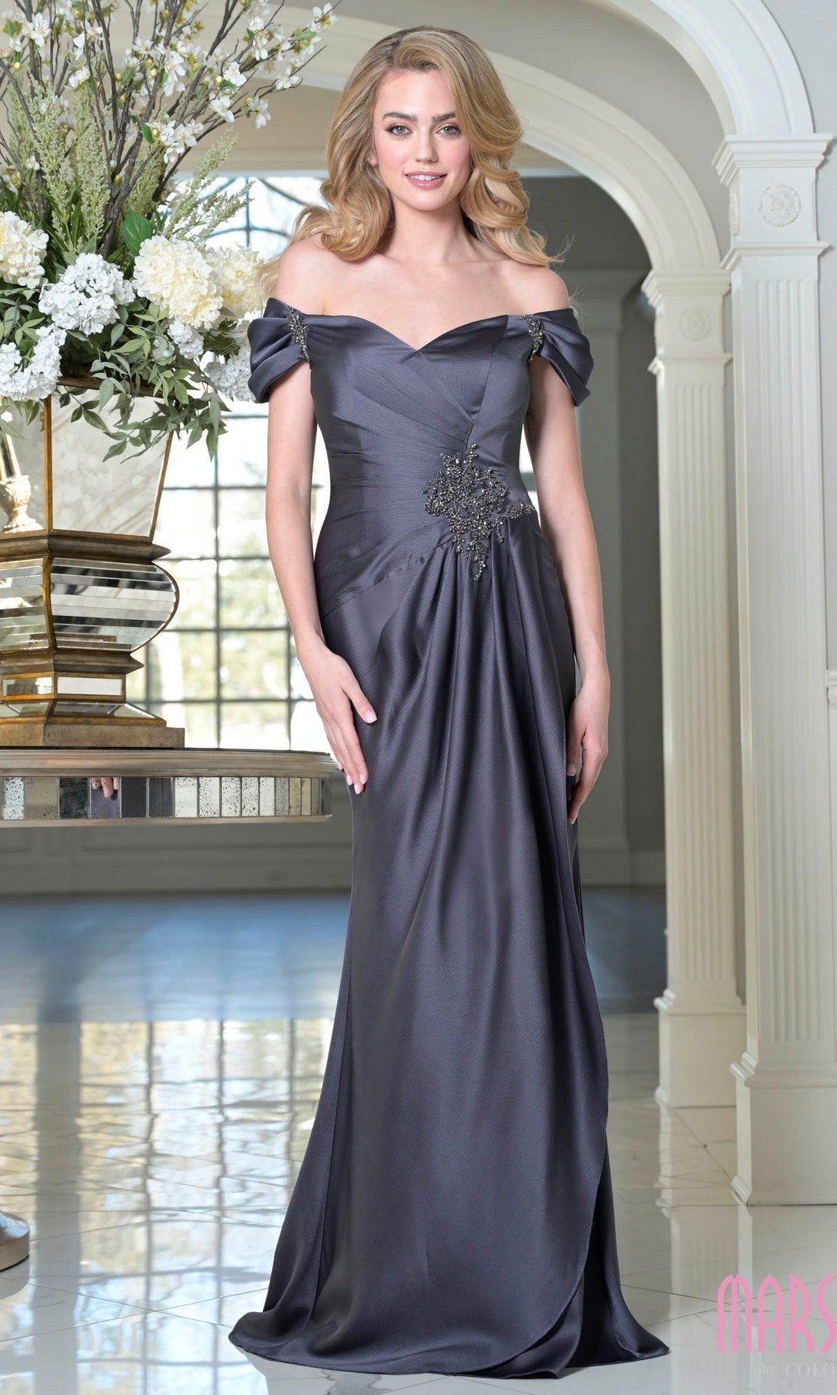 A woman stands indoors wearing the Off-Shoulder Long Formal Dress: Marsoni MV1351 in dark gray satin, featuring waist embellishments—a floor-length, elegant choice for special occasions.